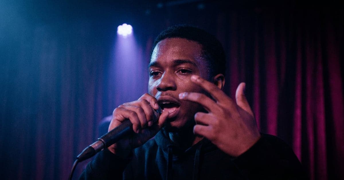 A rapper performing close to the camera under coloured stage light, mid-line.