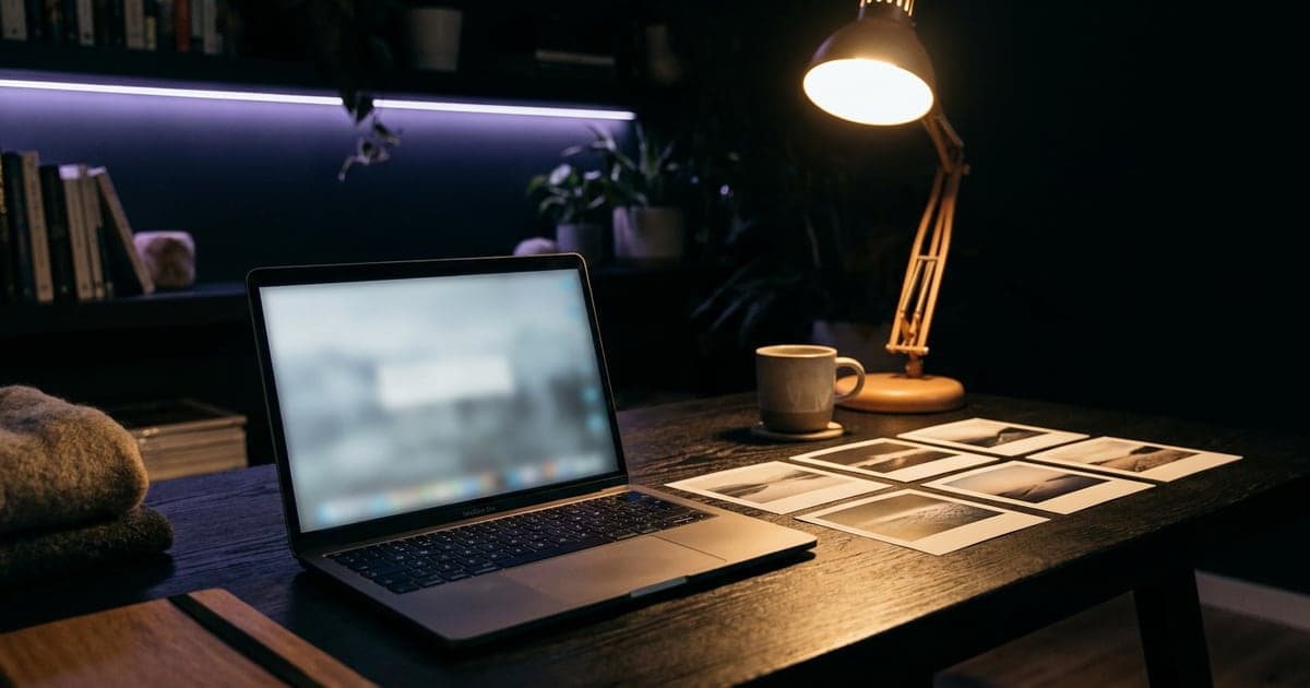 A laptop on a desk at night beside a row of freshly printed photographs, a desk lamp on.